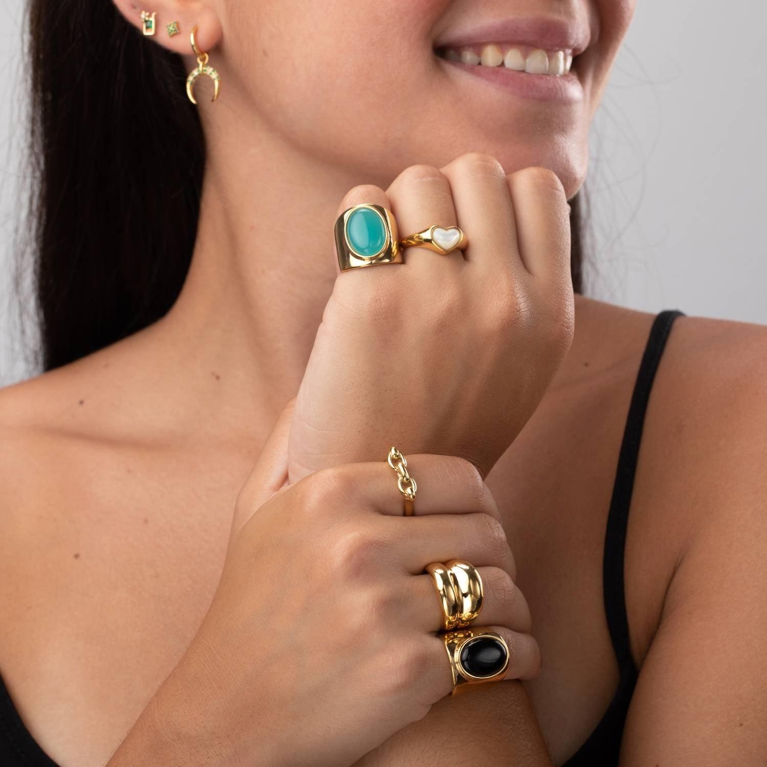 Mujer con anillos dorados de diseño exclusivo, incluyendo modelos con piedra verde, corazón nacarado y ónix negro, combinados con pendientes dorados en Cobalto Accesorios