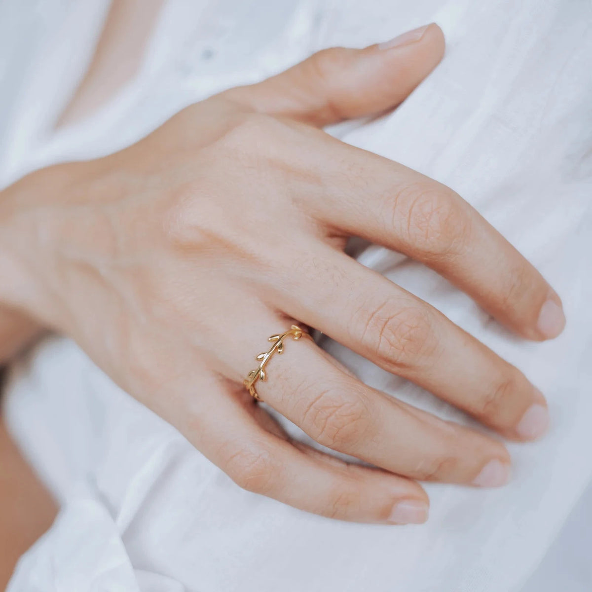 Detalle de un anillo dorado con diseño de rama y pequeñas hojas, colocado en la mano de una mujer sobre una blusa blanca.