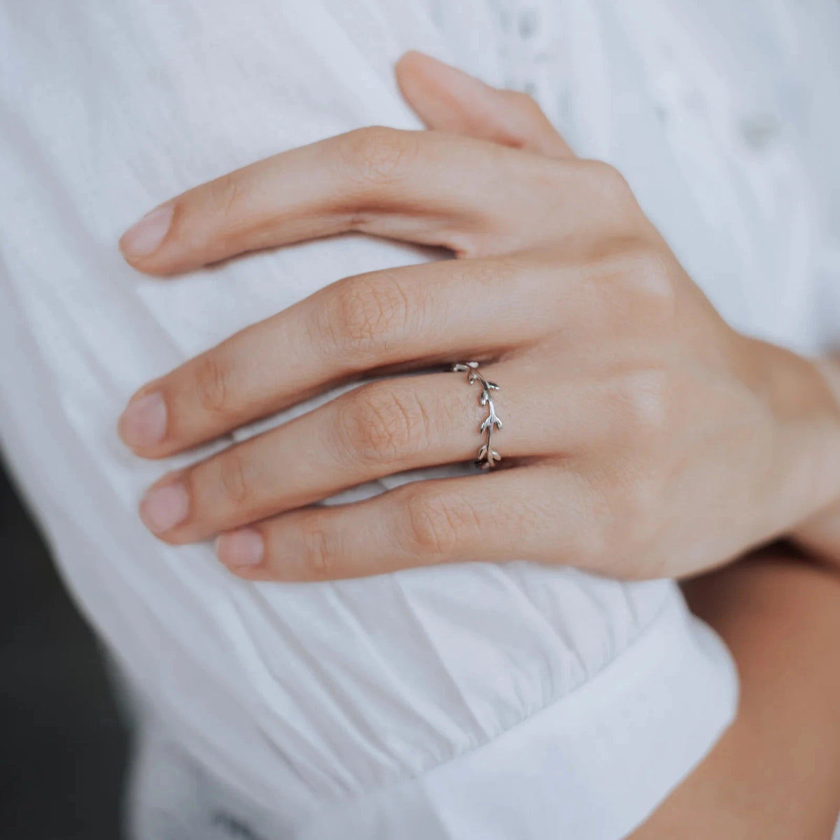Mano de una mujer apoyada sobre una blusa blanca, con un anillo plateado en forma de rama con pequeñas hojas.