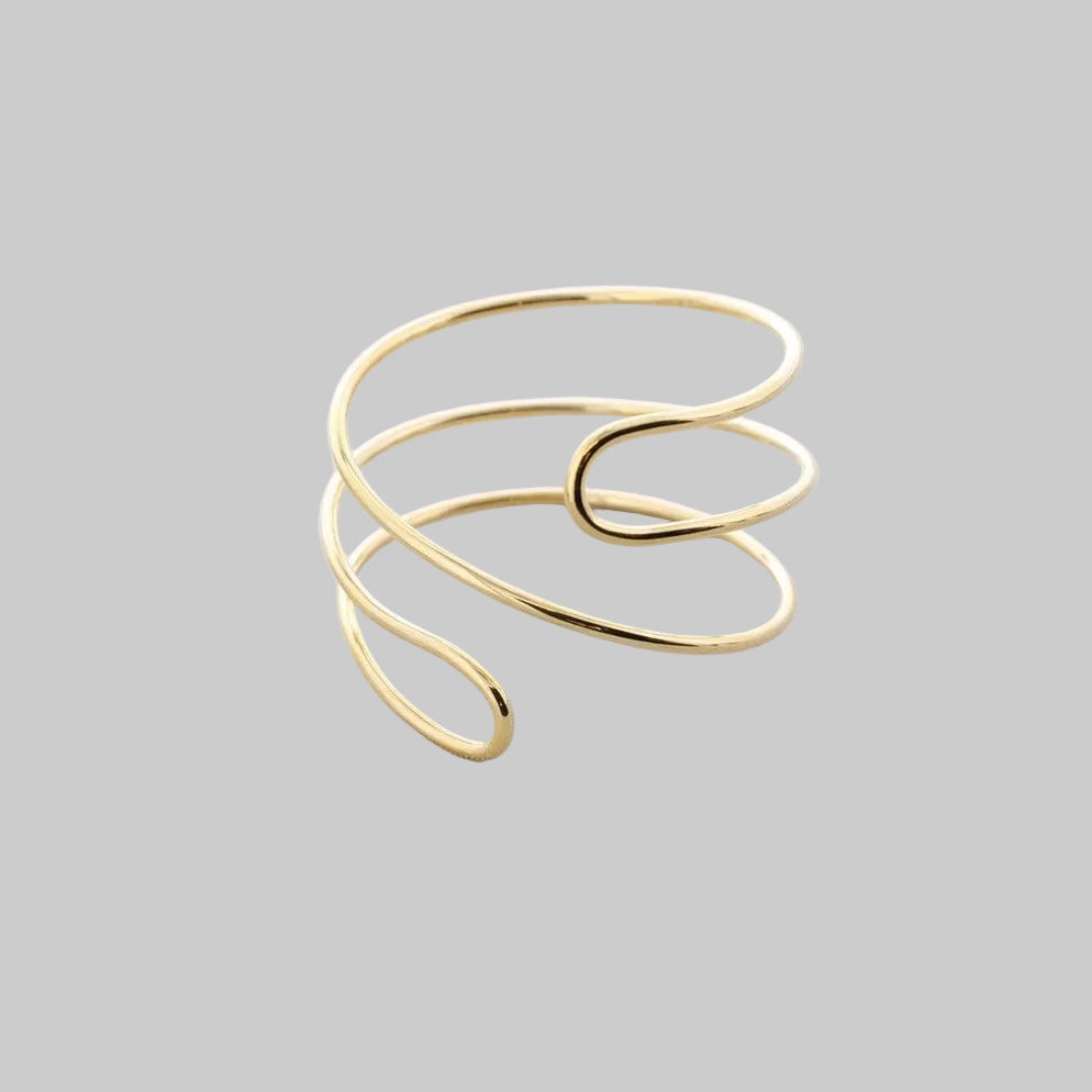 Brazalete de acero ajustable en acabado dorado con diseño minimal de líneas envolventes, fotografiado sobre fondo claro.