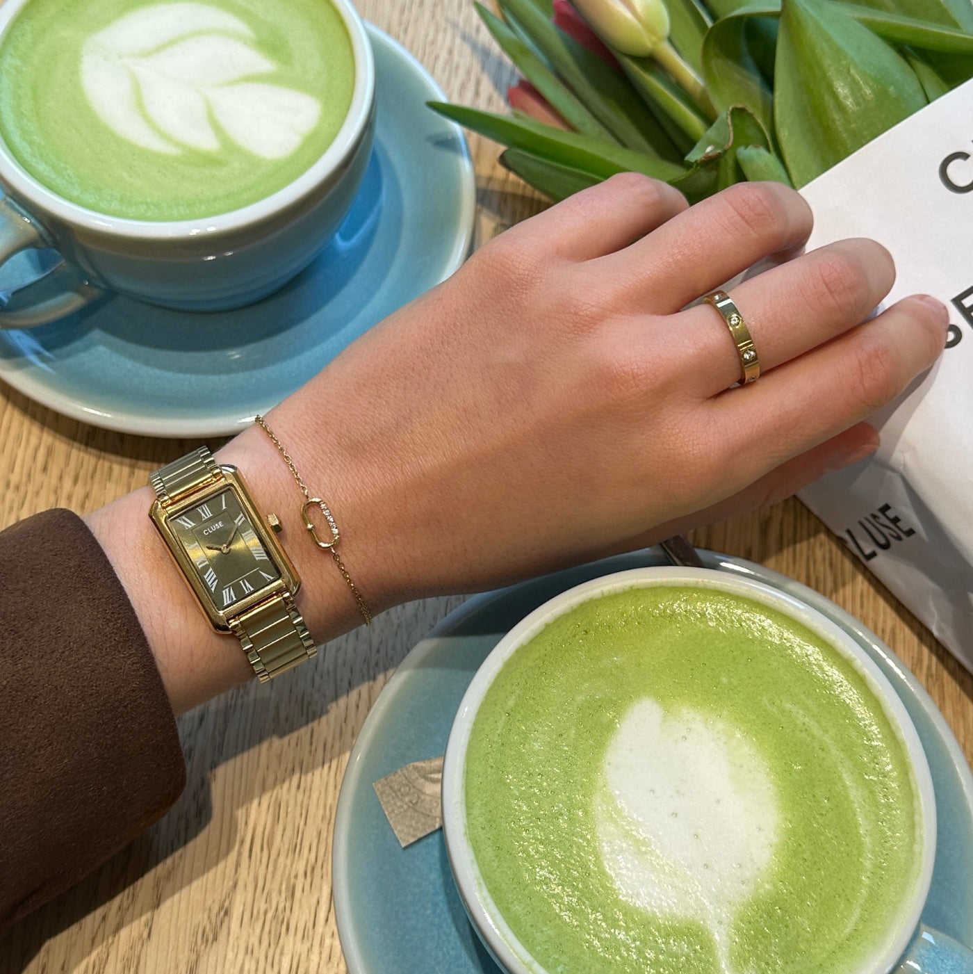 Reloj CLUSE Belisenna verde oliva y dorado en muñeca junto a café matcha, look casual y luminoso