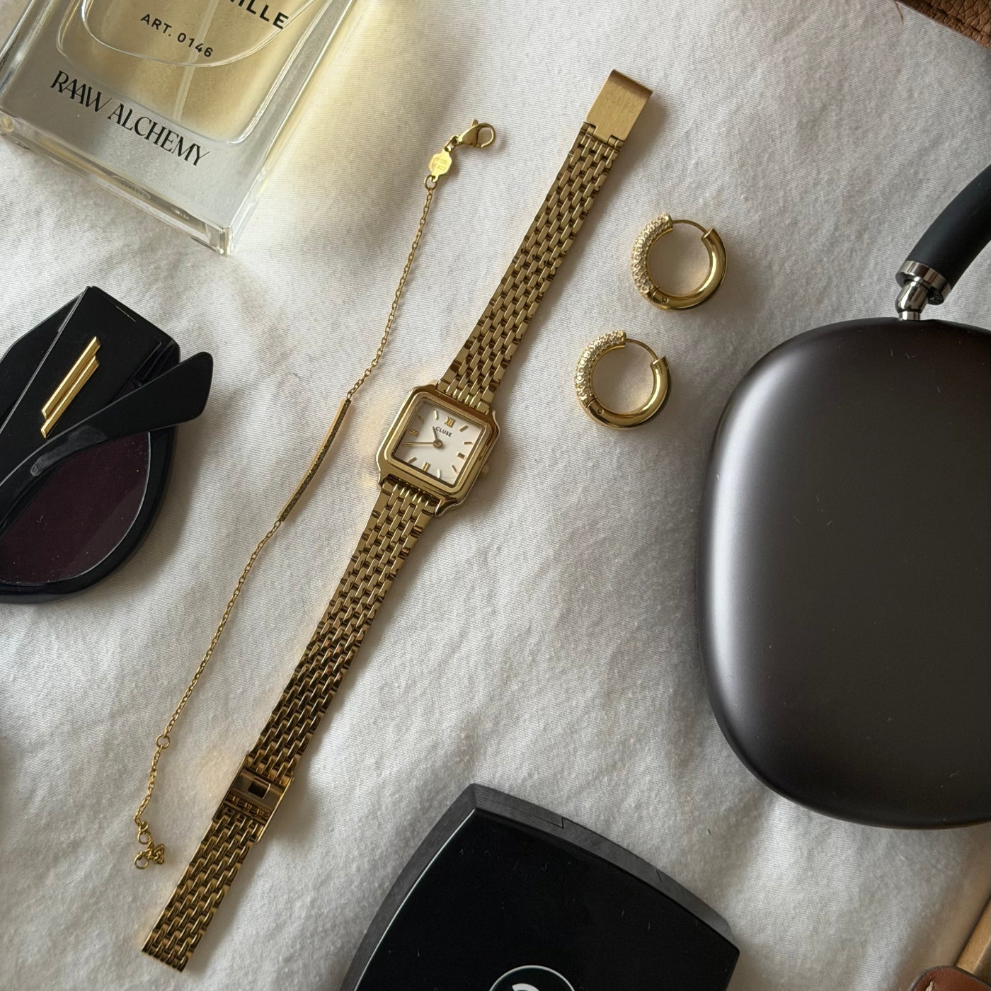 Reloj CLUSE Gracieuse Mini dorado con esfera blanca en flat lay junto a accesorios dorados, gafas y estuche sobre tela
