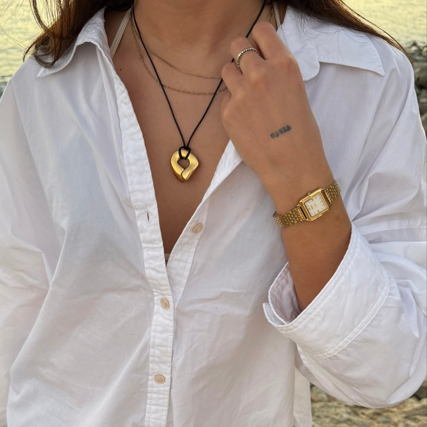 Reloj CLUSE Gracieuse Mini en acabado dorado y esfera blanca, combinado con camisa blanca y collares en una foto exterior