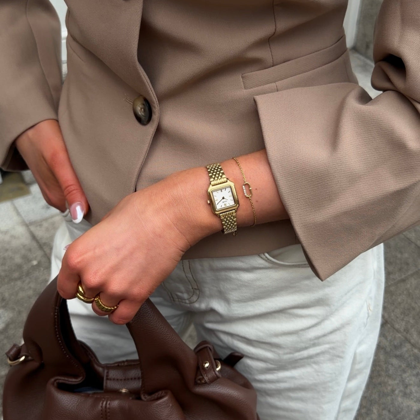 Reloj CLUSE Gracieuse Mini dorado con esfera blanca, puesto con blazer beige y bolso marrón en una escena urbana.