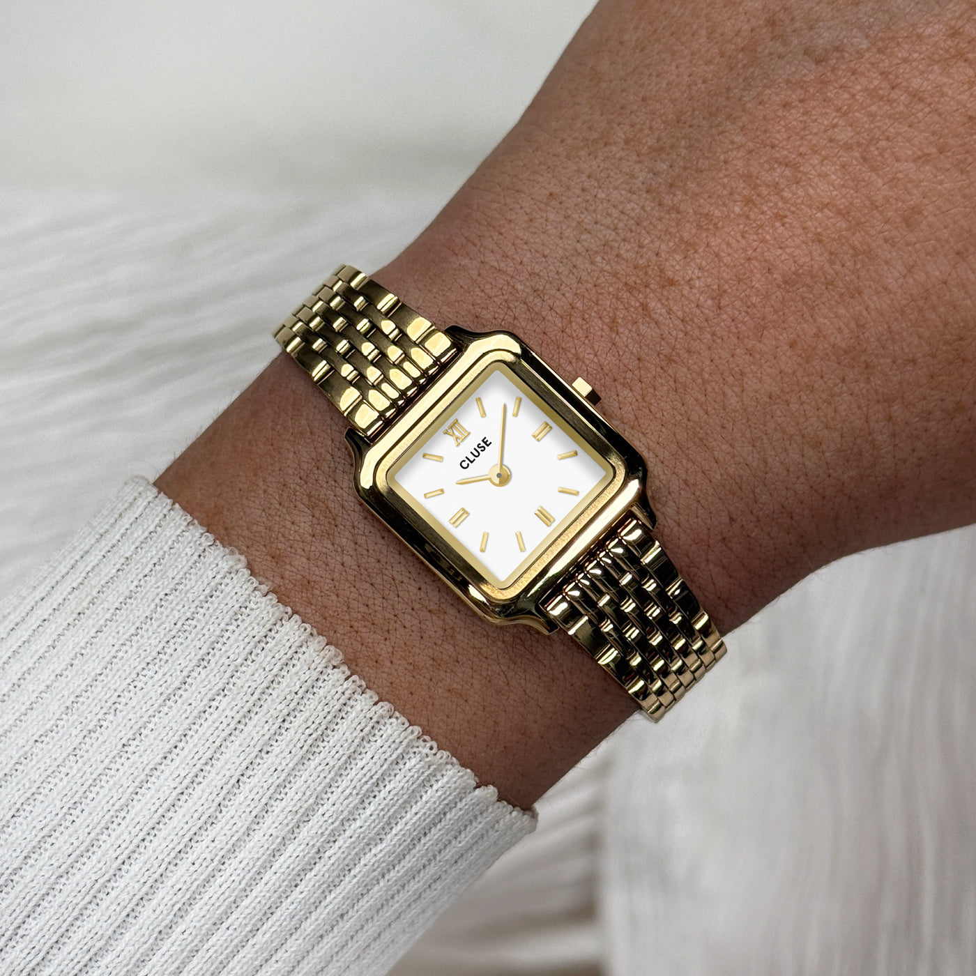 Reloj CLUSE Gracieuse Mini con esfera blanca y acabado dorado, fotografiado de cerca en la muñeca con luz suave.