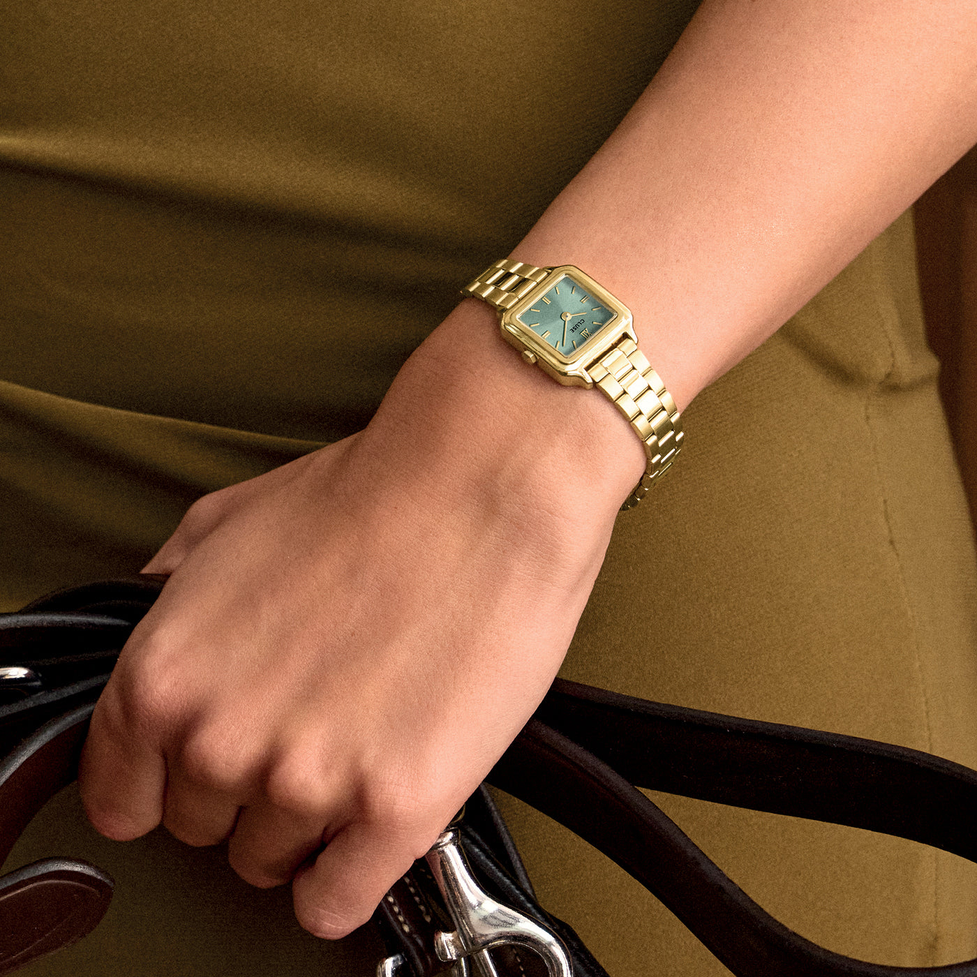 Reloj CLUSE Gracieuse Mini dorado con esfera verde salvia, puesto en la muñeca en una foto de estilo con ropa en tonos verdes.