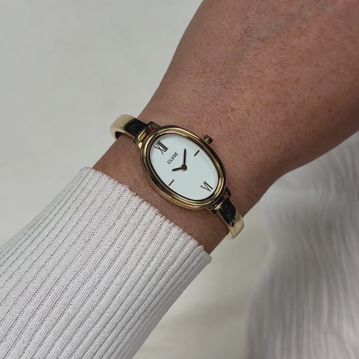Reloj Cluse L'Ovale en dorado y blanco visto de frente en muñeca sobre manga de tejido blanco