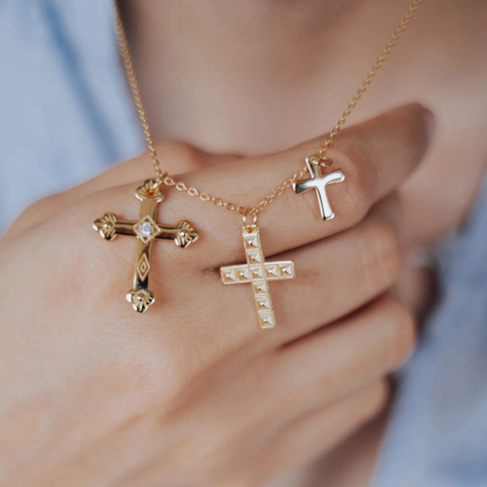 Collar de tres cruces llevado sobre camisa azul claro, con los colgantes sostenidos en la mano