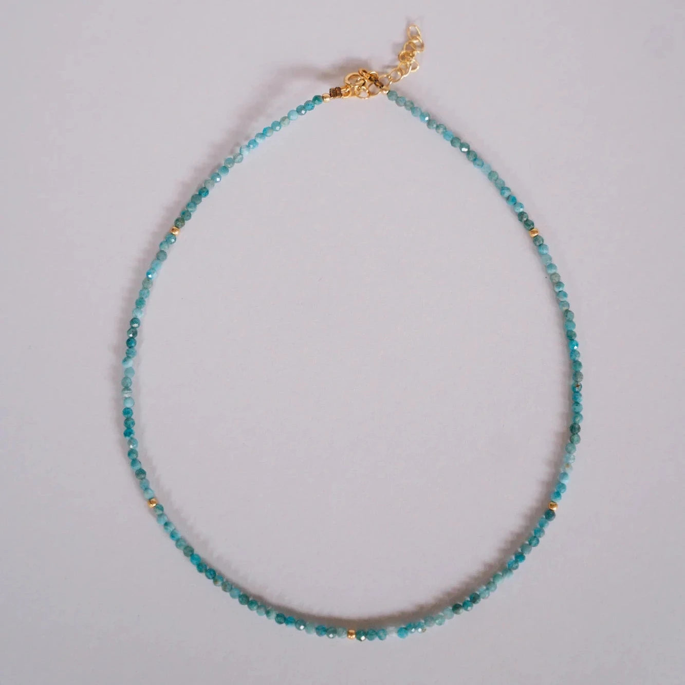 Collar de piedras facetadas neo-apatite azul verdoso con separadores dorados, vista cenital sobre fondo gris