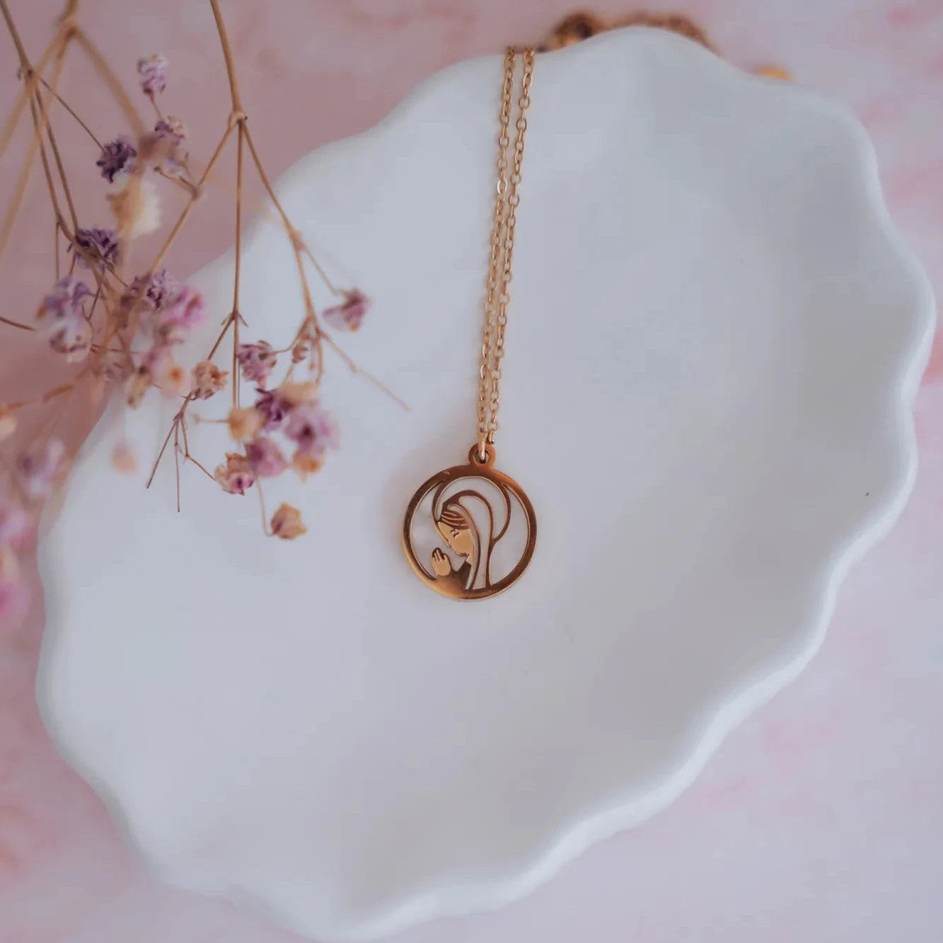 Collar de la Virgen Niña en acero dorado con nácar apoyado en un plato blanco con flores secas