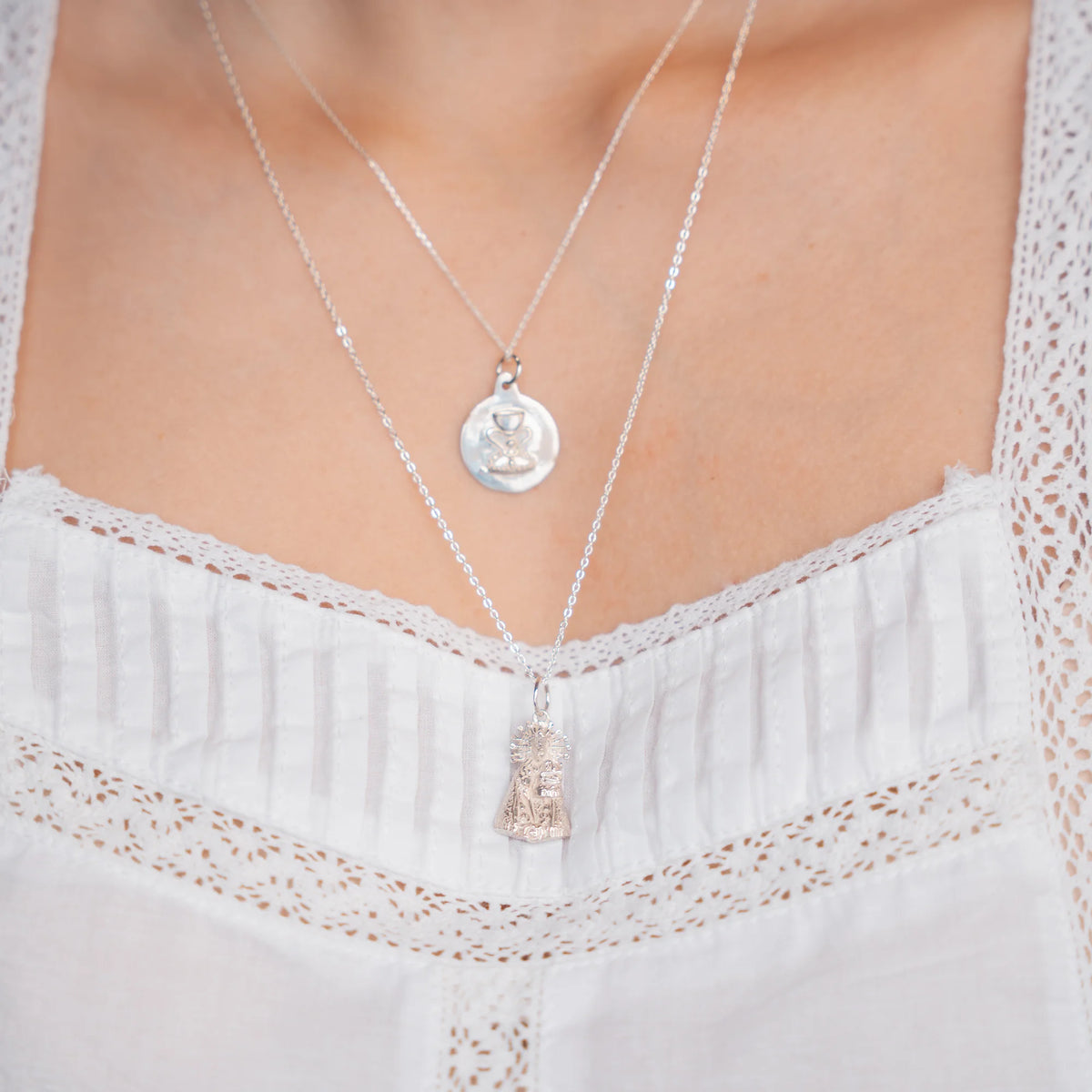 Figura de la Virgen de los Desamparados en plata puesta en cadena larga, bajo una medalla en cadena corta, sobre blusa blanca