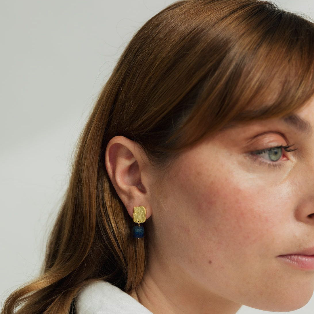 Pendiente Bruma puesto en la oreja, con pieza superior dorada texturizada y piedra lapislázuli azul, en retrato lateral.