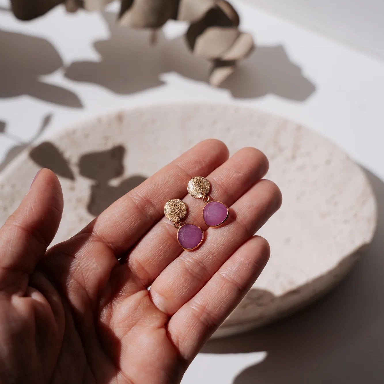 Mano sujetando dos pendientes con chapita dorada y piedra natural colgando.