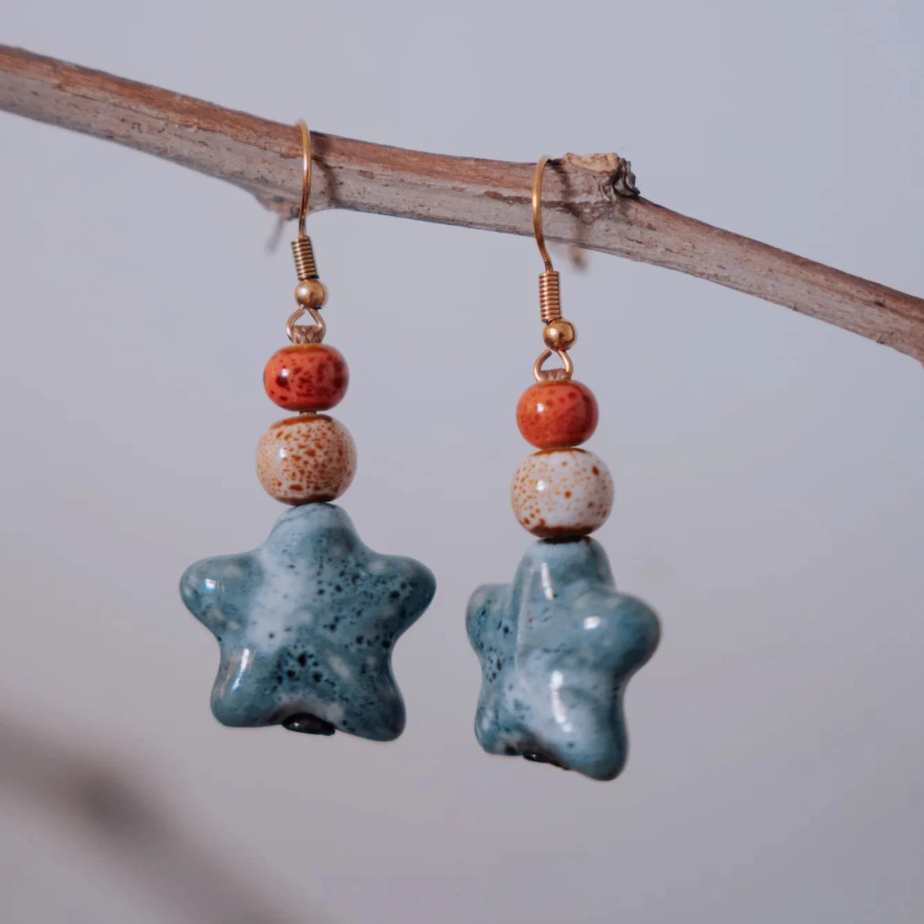 Pendientes estrella de cerámica azul teal con cuentas naranja y beige moteadas colgando de rama natural