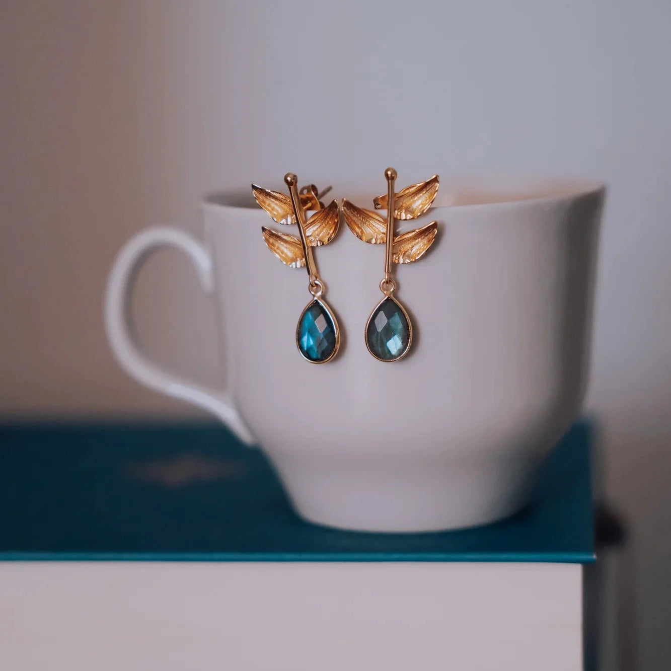 Pendientes Aurora Labradorita apoyados en taza blanca sobre libro azul, detalle lifestyle