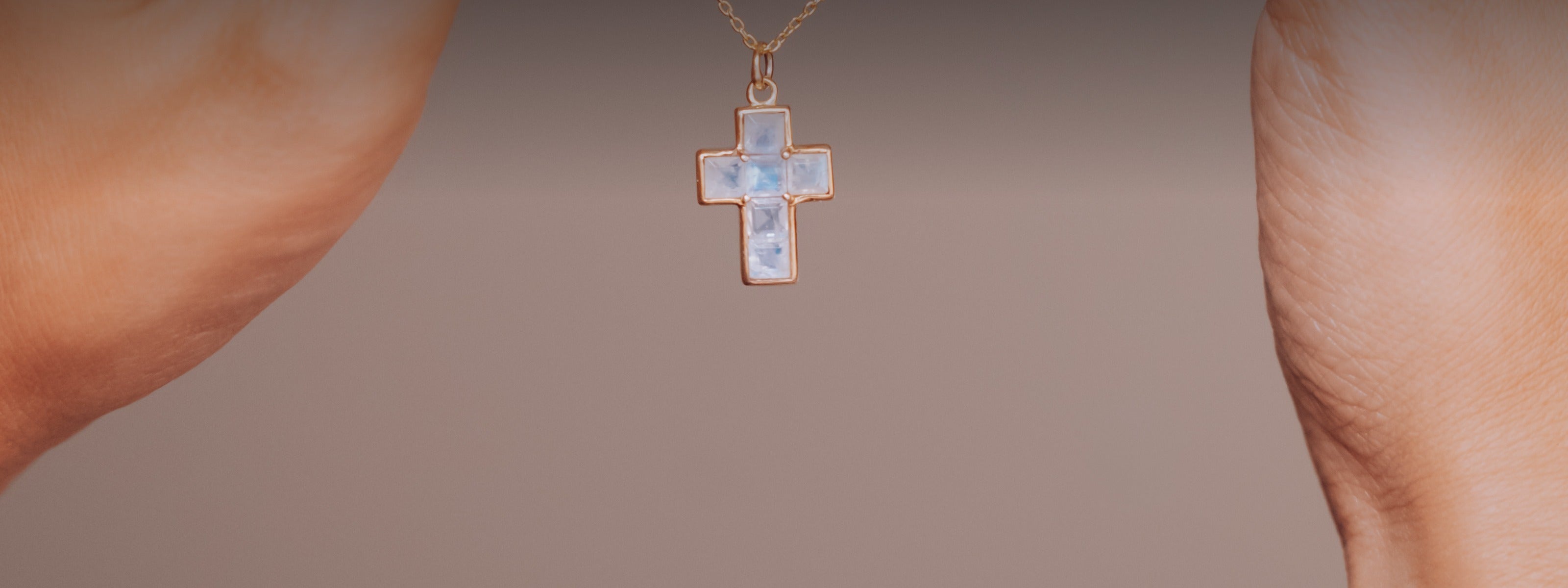Dos manos sosteniendo un collar de cadena dorada con una cruz de piedra luna, sobre fondo neutro