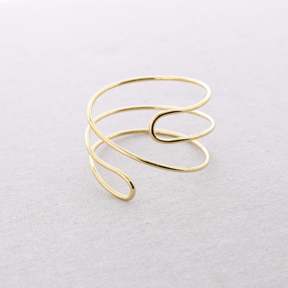 Brazalete de acero ajustable en acabado dorado con diseño minimal de líneas envolventes, fotografiado sobre fondo claro.
