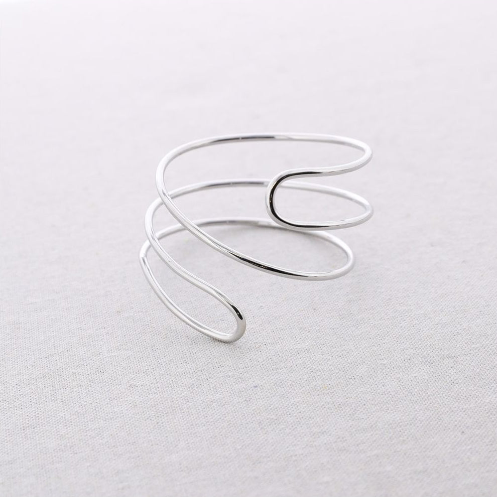 Brazalete de acero ajustable en acabado plateado con diseño minimal de líneas envolventes, fotografiado sobre fondo claro.
