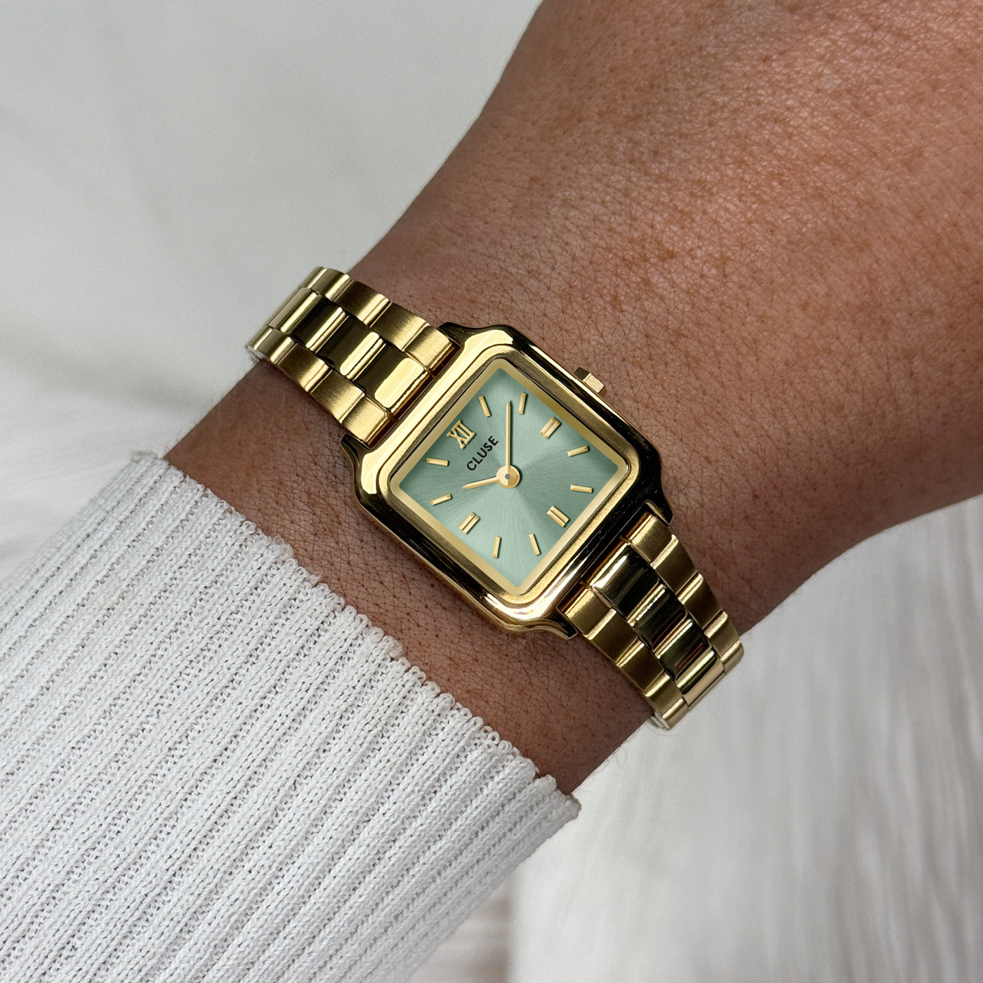 Reloj CLUSE Gracieuse Mini en acabado dorado con esfera verde salvia, puesto en la muñeca con jersey claro en una escena luminosa
