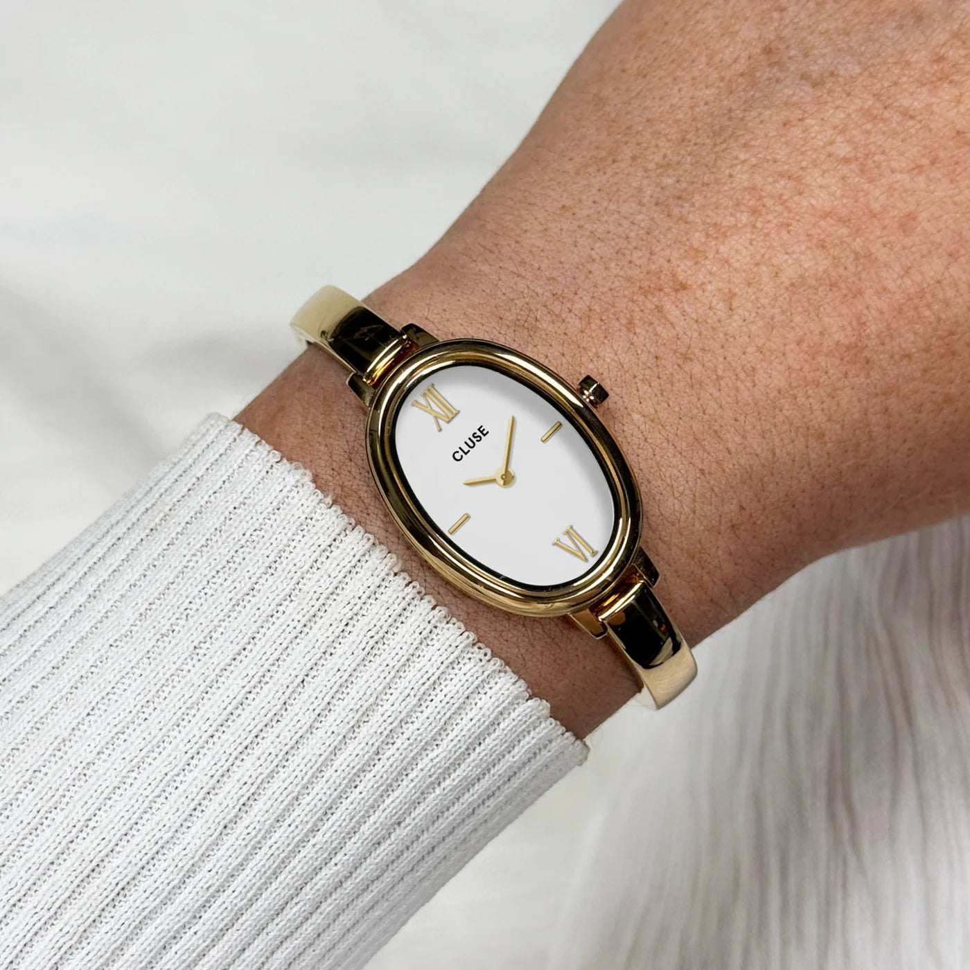 Reloj Cluse L'Ovale dorado y blanco en muñeca sobre jersey de punto blanco