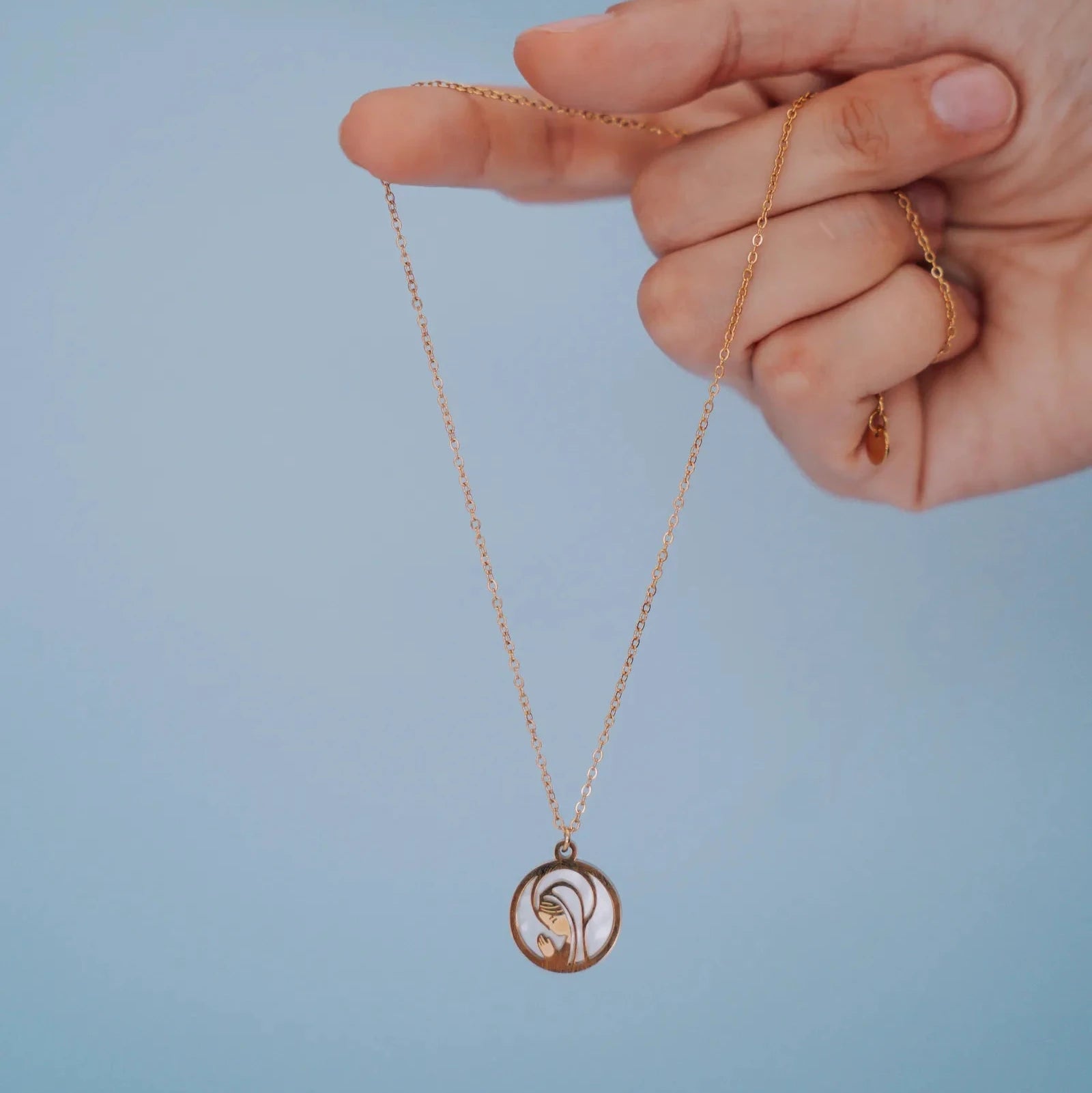 Colgante de la Virgen Niña en acero dorado con nácar sostenido con la mano sobre fondo azul