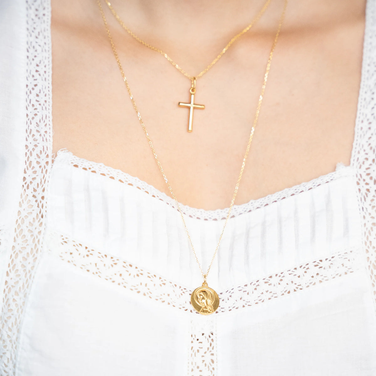 Medalla de la Virgen Niña dorada puesta en cadena larga, con una cruz palo en cadena corta, sobre blusa blanca