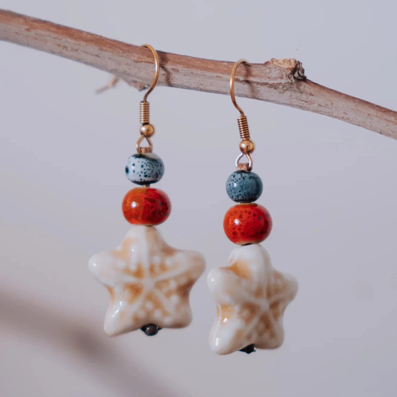 Pendientes estrella de cerámica blanca con cuentas cerámicas naranja y azul colgando de rama natural